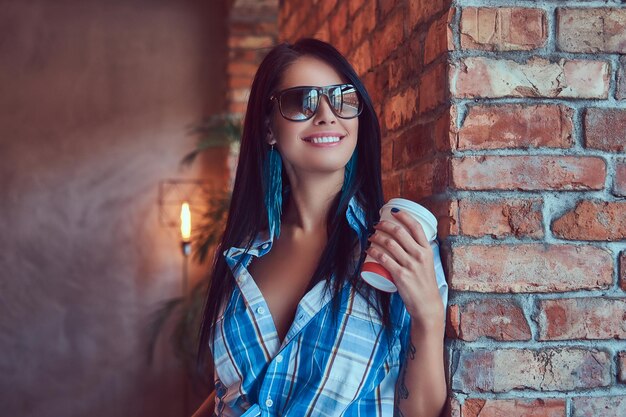 A happy sexy brunette in flannel shirt and sunglasses holds a cup of coffee posing against a brick wall.