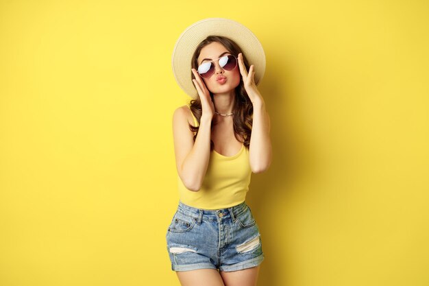 Счастливая сексуальная молодая женщина в отпуске, в соломенной шляпе, солнцезащитных очках, смеется и улыбается, стоя на желтом фоне.