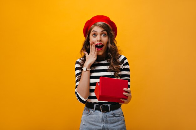 Happy shocked woman in red beret holds gift box. Surprised lady with wavy hair in bright hat and striped shirt rejoices.