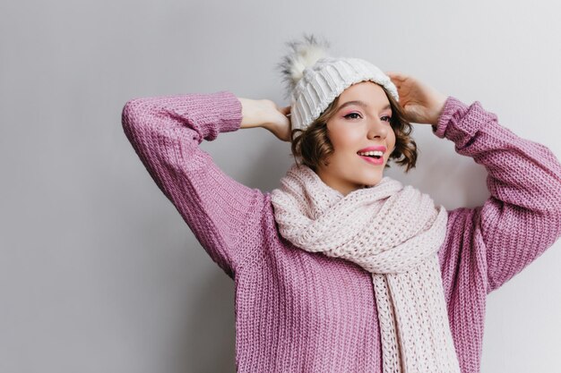 Happy short-haired female model posing with hands up on light background. Studio shot of spectacular brunette girl wears knitted winter accessories.
