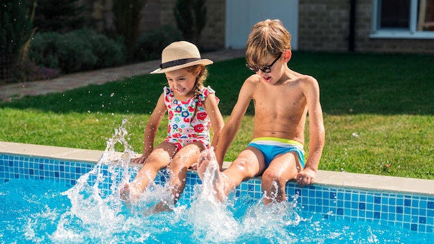 Free photo happy siblings are sitting on the side of the pool and splashing their feet in the water