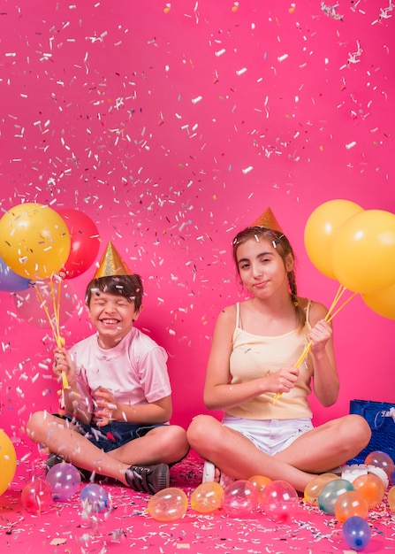 Happy siblings enjoying party with balloons and confetti over pink background