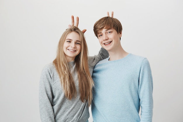 Happy siblings smiling. Girl and boy with braces make bunny ears up behind