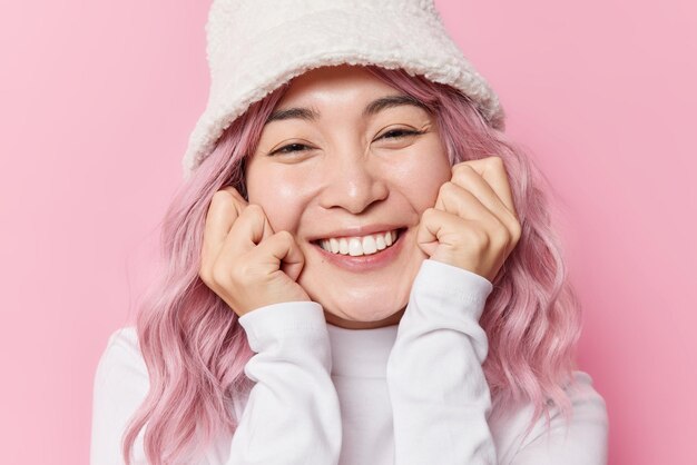 Happy sincere Asian woman keeps hands on cheeks smiles pleasantly shows white teeth wears fashionable panama and white turtleneck feels very glad isolated over pink background Emotions concept