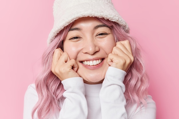 무료 사진 행복한 성실한 아시아 여성이 뺨에 손을 얹고 미소를 지으며 하얀 치아가 세련된 파나마를 입고 흰색 터틀넥은 분홍색 배경 감정 개념에 고립되어 매우 기쁘게 생각합니다.