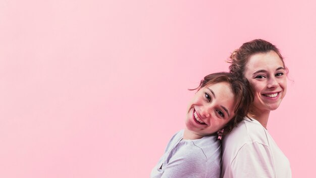 Happy sisters sitting back to back against pink background