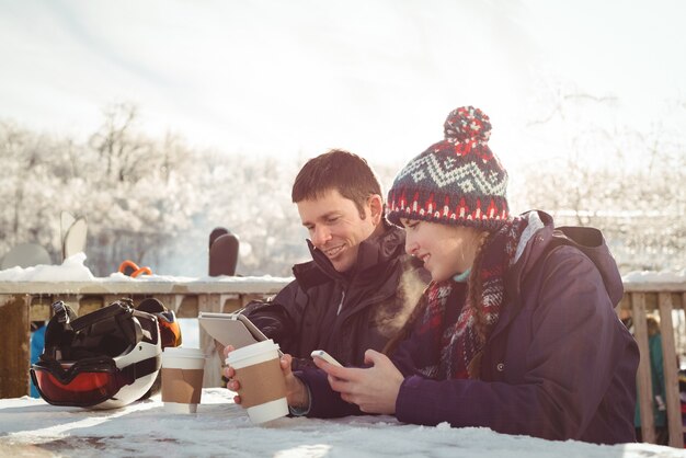 携帯電話とデジタルタブレットを使用しながらテーブルに座って幸せなスキーヤーのカップル