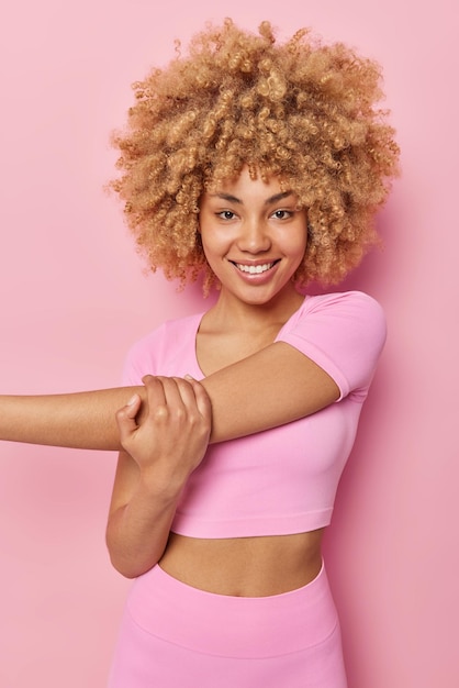 Бесплатное фото Счастливая стройная молодая женщина с вьющимися волосами протягивает руки перед тренировкой, разогревается, остается в хорошей физической форме, радостно улыбается, носит топ и леггинсы, изолированные на розовом фоне, упражнения в помещении