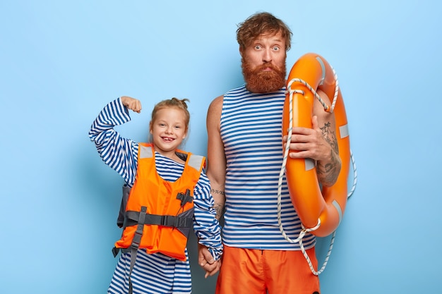 Happy small child in orange lifejacket raises arm and shows muscle holding hands with father