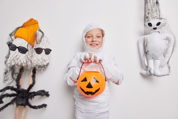 Happy small halloween child plays trick or treat jack o lantern pumpkin wrapped in white fabric surrounded by holiday attributes isolated on white 