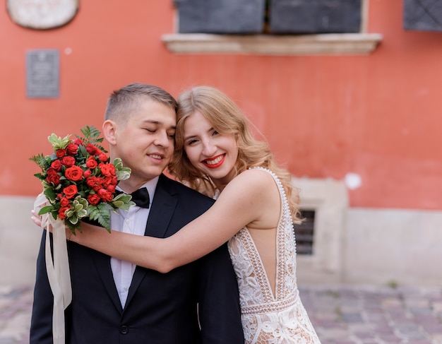 Foto gratuita la coppia di sposi sorrisa felice sta abbracciando davanti alla parete rossa all'aperto, il giorno delle nozze, matrimonio ufficiale