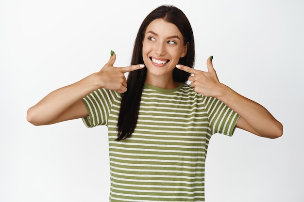 Happy smiling adult woman pointing fingers at her smile showing white healthy teeth standing over white background