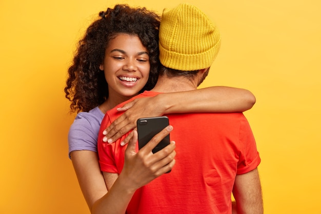 Happy smiling Afro American woman embraces boyfriend who stand back at camera, holds cell phone