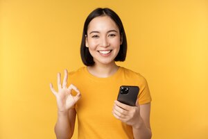 happy smiling asian girl holding mobile phone and showing okay recommending application on smartphone standing over yellow background