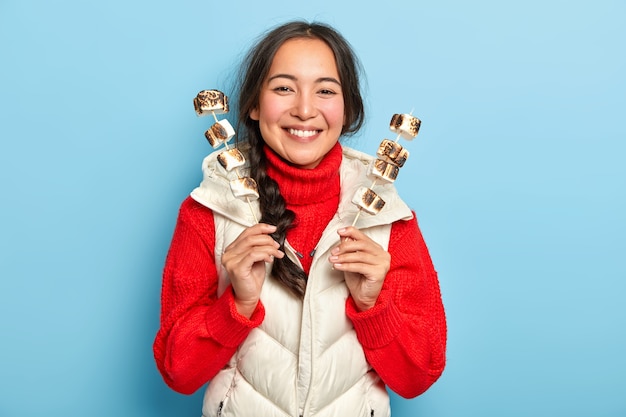 La ragazza asiatica sorridente felice tiene i bastoni con marshmallow arrosto deliziosi aromatici, gode del picnic in campagna, indossa vestiti caldi