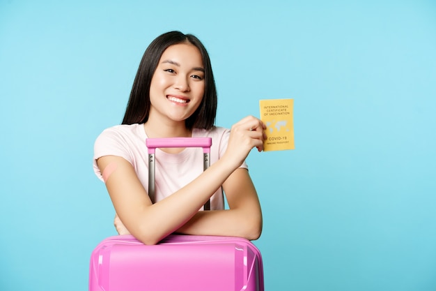 Счастливая улыбающаяся азиатская туристка стоит с симпатичным чемоданом в аэропорту и показывает международную вакцину против ...
