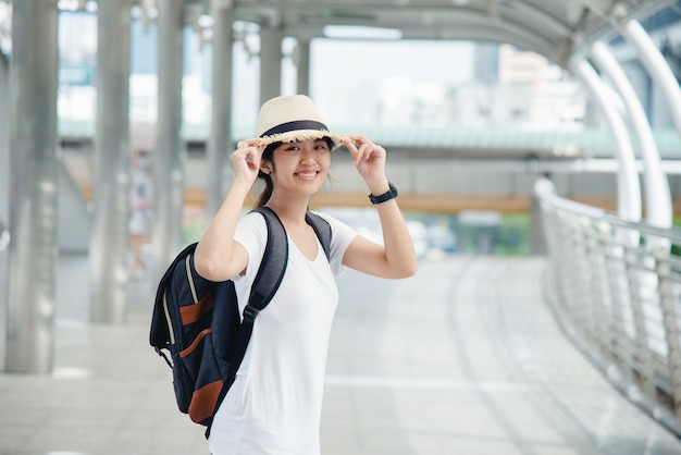 Счастливая усмехаясь азиатская девушка студента с рюкзаком на предпосылке города