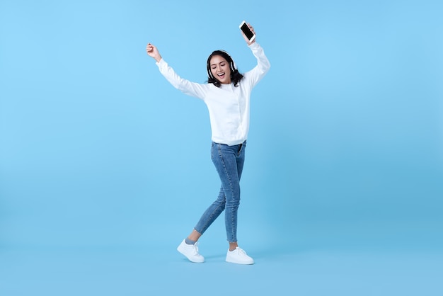 Happy smiling Asian woman wearing wireless headphones listening to music and dancing on blue.