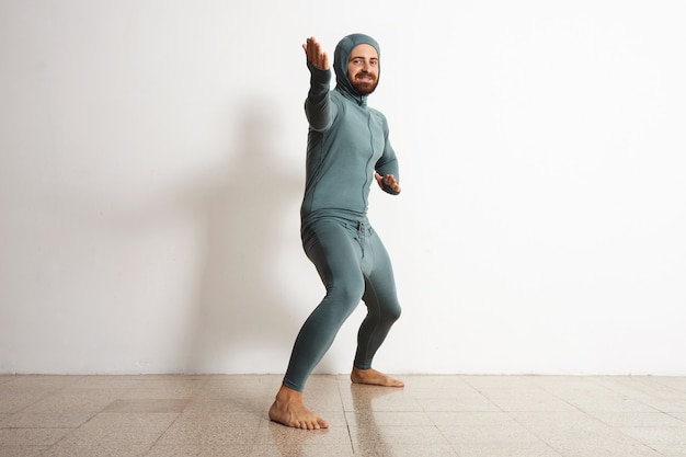 Happy smiling bearded fitted male wearing snowboarding thermal baselayer suite from merino wool and acts like a ninja in welcome position, isolated on white