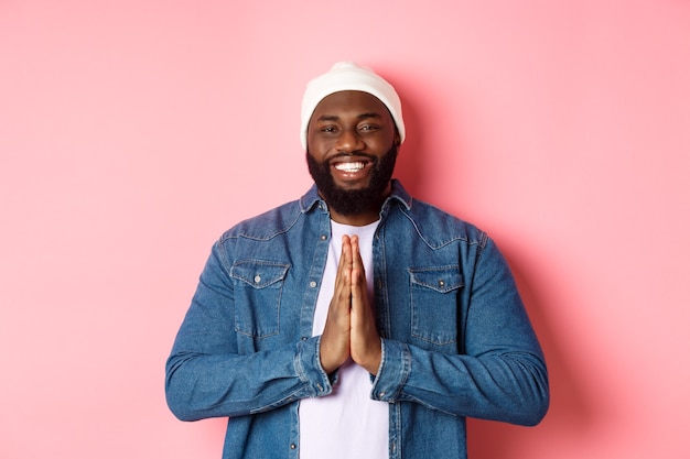 Happy smiling Black man saying thank you, holding hands in pray or namaste gesture, standing grateful against pink background