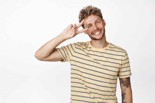 Happy smiling blond man shows peace sign salute and looking at camera positive standing over white background
