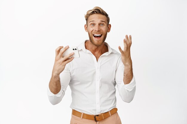 흰색 배경 복사 공간 위에 서 있는 스마트폰에서 비디오를 보고 있는 휴대전화에서 이기고 행복한 미소 짓는 금발 남자