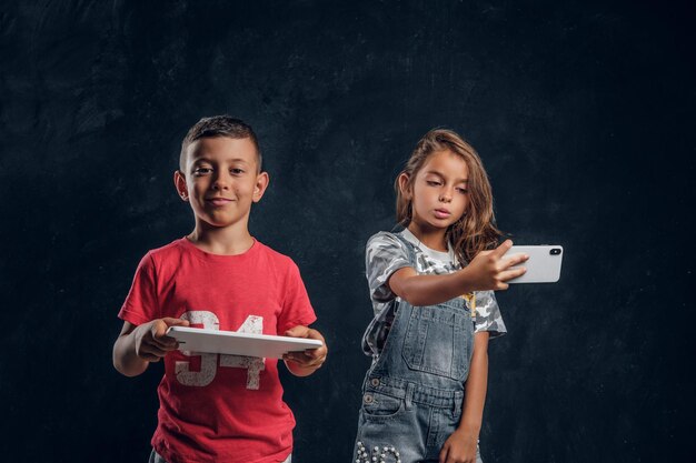 Happy smiling boy is holding tablet while girl is making selfie by mobile phone.