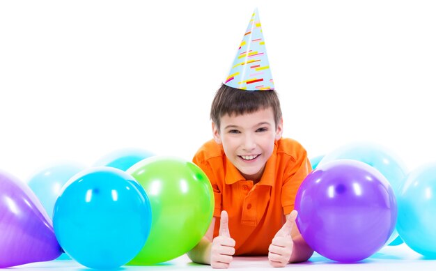 Счастливый улыбающийся мальчик в оранжевой футболке, лежащий на полу с разноцветными воздушными шарами и показывающий большие пальцы руки вверх - изолированный на белом