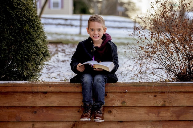 Happy smiling boy sitting on a wooden fence and reading a book in a park