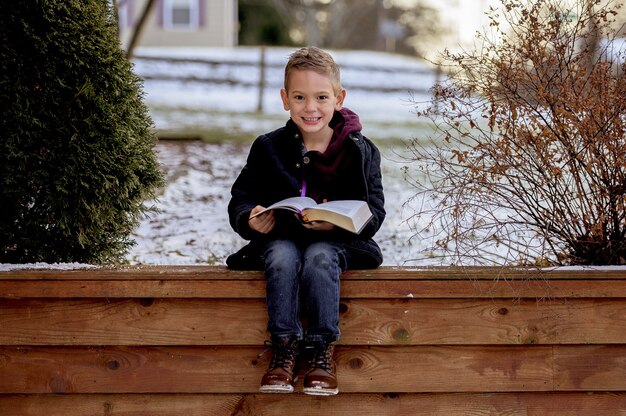 Happy smiling boy sitting on a wooden fence and reading a book in a park