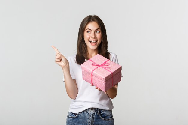 Happy smiling brunette girl holding birthday gift and pointing finger left at logo.