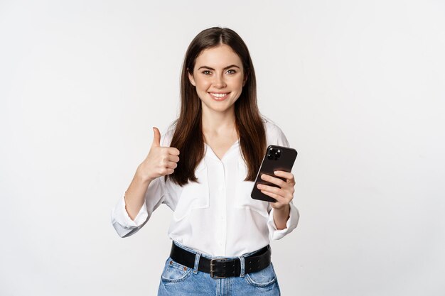 Happy smiling corporate woman female model showing thumb up holding smartphone using cellphone stand...