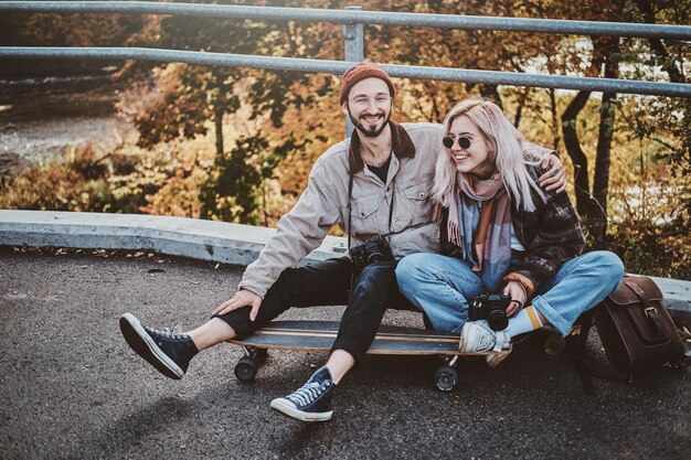 Happy smiling couple is chilling on longboard with golden trees at background.