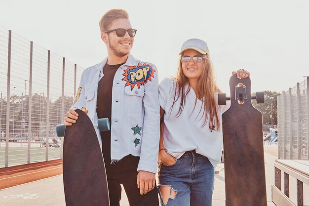 행복한 미소를 짓고 있는 커플이 롱보드를 들고 여름 거리에 서 있습니다. 그들은 선글라스를 착용합니다.