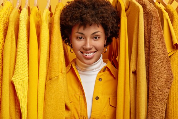 Happy smiling curly haired woman searches what to wear, stands between bright outfits