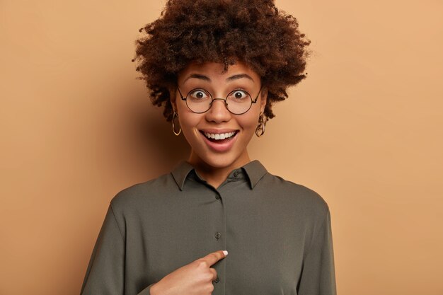 행복하게 웃고있는 곱슬 곱슬 한 여자는 누군가가 그녀의 이름을 언급하고 자신을 가리키며 놀랐고,이기거나 선택 될 것이라고 예상하지 않았고, 상사에게 그녀가 정확히 승진했는지 묻고 둥근 안경과 셔츠를 입었습니다.