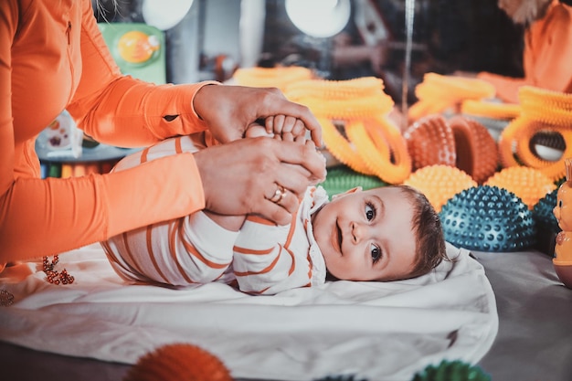 Happy smiling cute toddler is lying on the special table surrounded by ortopedic toys while expirienced masseur making massage for him.