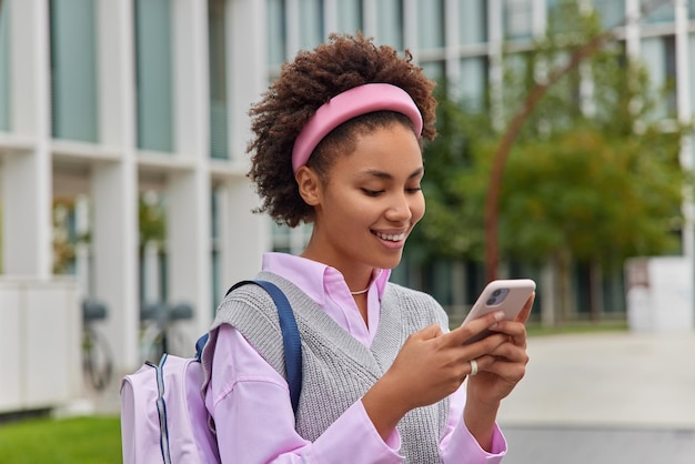 Happy smiling female student wears headband shirt and vest poses with backpack sends text messages on her way to university