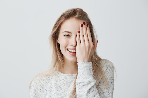 Счастливая улыбающаяся женщина с привлекательной внешностью и светлыми волосами в свободном свитере с широкой улыбкой и хорошим настроением закрывает глаза рукой