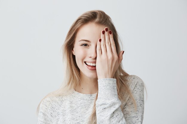 Happy smiling female with attractive appearance and blonde hair wearing loose sweater showing her broad smile having good mood closing her eye with hand