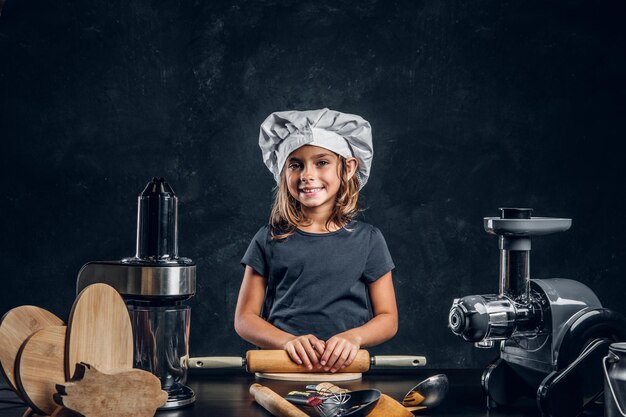 Happy smiling girl in chef's hat is posing for photographer with kitchen equipment.