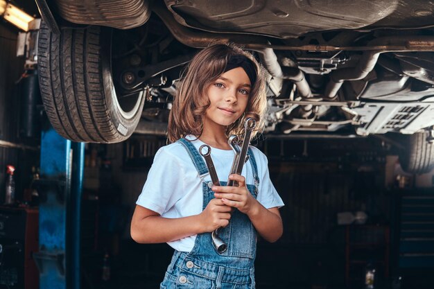 행복한 미소 짓는 소녀가 렌치를 손에 들고 자동차 작업장에서 차 아래에 서 있습니다.