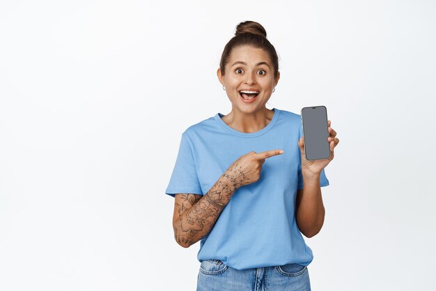 Happy smiling girl pointing finger at mobile phone, showing interface, empty display, standing in blue t-shirt against white background.