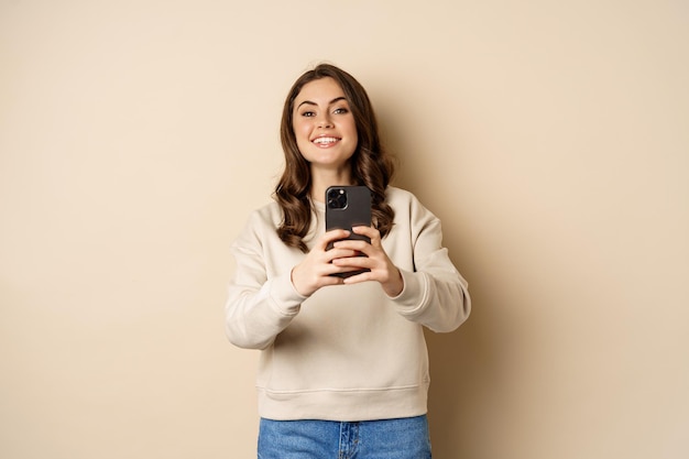 Happy smiling girl recording video, taking picture on smartphone and looking at camera, standing over beige background