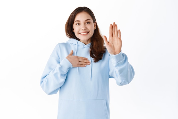 Happy smiling girl student raise her hand and touching chest, volunteering, making me gesture, talking about herself, standing against white wall