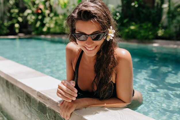 Счастливая улыбающаяся девушка с темными влажными волосами в купальнике, купающемся в бассейне в солнечный теплый летний день. Фото высокого качества