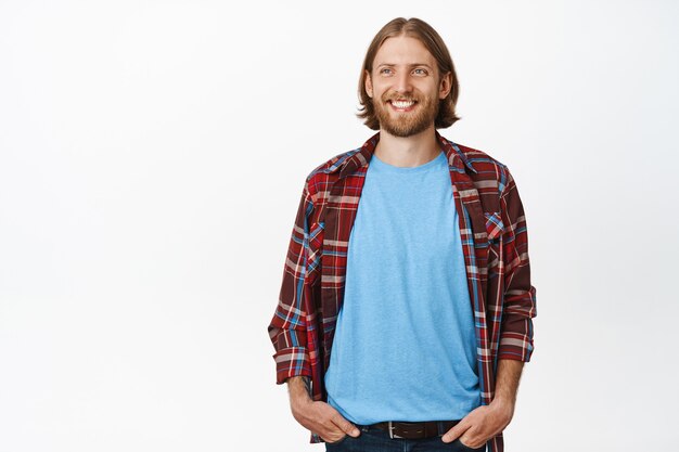 Happy smiling hipster guy, blond adult man with beard, looking hopeful and cheerful aside, standing casual with hands in pockets of jeans on white