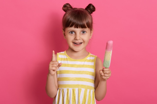 아이스크림을 들고 흰색과 노란색 줄무늬가있는 여름 드레스를 입고 그녀의 검지 손가락으로 가리키는 행복 웃는 아이 소녀.