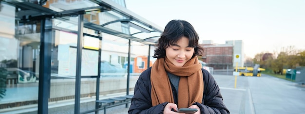 無料写真 バス停に立ってスマートフォンを見て携帯電話を使用して幸せな笑顔の韓国の女の子