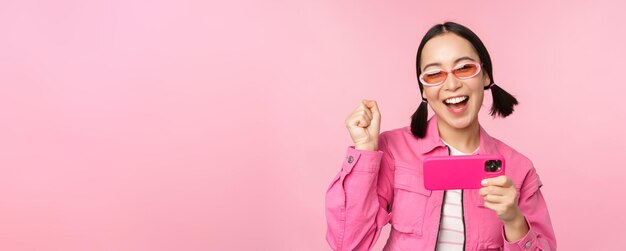 Happy smiling korean girl winning on mobile phone looking at horizontal smartphone screen and rejoicing achieve goal celebrating standing over pink background